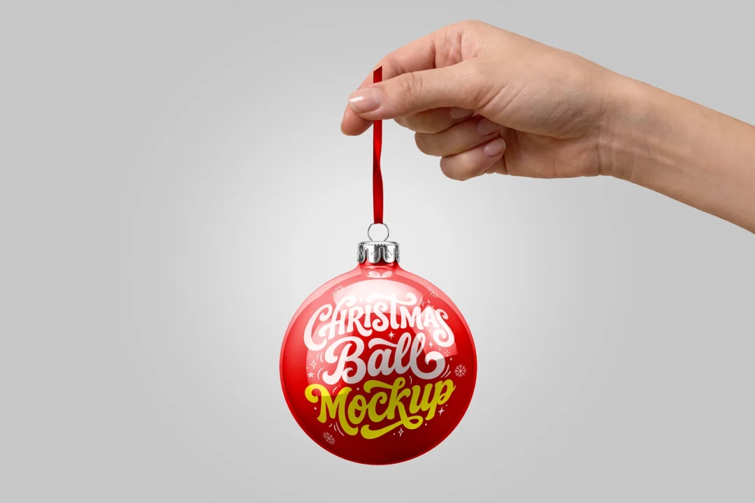 Christmas ball mockup in hand, glossy red bauble with silver cap hanging from a red ribbon on gray
